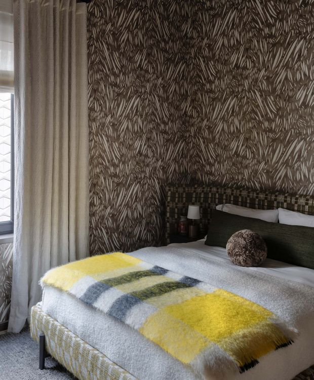 A cozy bedroom corner featuring a neatly made bed with a bold yellow, grey, cream and olive checked throw, dark green cushions, a plaid upholstered headboard, and a small bedside lamp. The walls are covered in a dramatic dark botanical-patterned wallpaper, while full-height textured curtains frame a window on the left. The room has a layered, richly patterned and tactile interior styling.