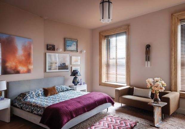 A warm, traditional bedroom with a large upholstered bed, patterned blue-and-white bedding, and a deep burgundy throw. Blush-taupe walls are decorated with framed artwork, while timber-framed windows with timber Venetian blinds bring in natural light. A compact upholstered armchair and small side table sit beside the windows, with layered rugs and decorative lighting completing the room.