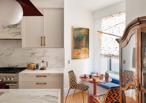 A warm, characterful kitchen and compact dining nook. The kitchen features pale fixed cabinetry with dark pulls, a dramatic white-and-grey marble backsplash and benchtops, a freestanding range, and a partial island. Beside it is a window seat dining area with a small round red table, patterned upholstered chairs, a vintage-style poster, and a large carved timber mirrored cabinet. A sculptural dark red rangehood and globe light add bold accents.