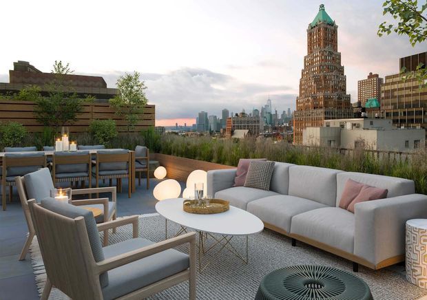 A sophisticated rooftop terrace lounge overlooking a dense urban skyline. The space features a large light-grey sectional sofa, woven lounge chairs, a white oval coffee table, a dining table with chairs, outdoor lighting, planters and tall grasses. The terrace has a calm, contemporary hospitality-like atmosphere, with a prominent historic high-rise tower and modern city buildings visible beyond.