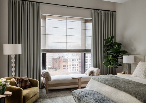 A softly lit, refined bedroom with a large city-facing window, full-height grey curtains, a built-in or freestanding window seat, upholstered bed, textured grey throw, bedside lamps, and an olive-green armchair. The neutral palette and layered textiles create a calm, luxurious atmosphere.
