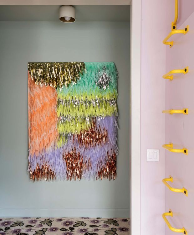 A vibrant, playful interior vignette featuring a pastel blue-green recessed wall niche with a large multicoloured shaggy textile artwork. The adjacent pale pink wall has a vertical row of bright yellow sculptural coat hooks, with white trim and a simple ceiling spotlight. A patterned floral carpet adds further colour and texture.