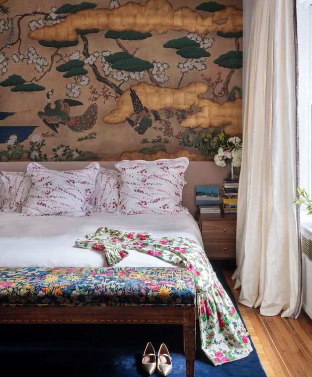A richly layered bedroom with a large traditional Japanese-style mural depicting birds, flowering branches, trees and clouds behind the bed. The bed is dressed in white linen with patterned floral pillows and a colourful floral throw, while a dark upholstered bench sits at the foot. Timber floorboards, a deep blue rug, bedside storage, books and a vase of flowers complete the warm, eclectic composition. Full-height white curtains frame a bright window on the right.
