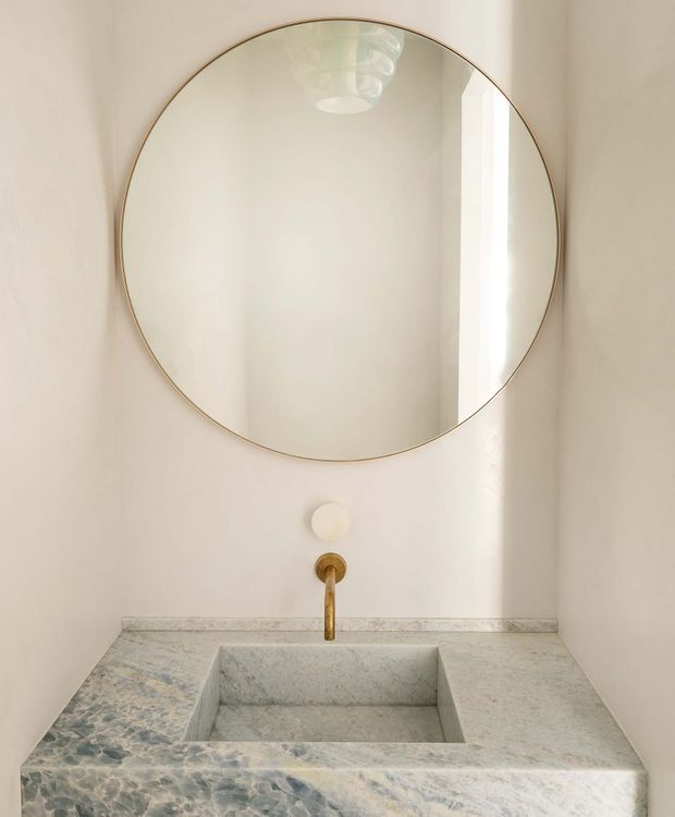 A refined bathroom vanity with a large round frameless-style mirror edged in a thin brass-gold frame, a wall-mounted brass tap, and a square integrated stone basin. The vanity and backsplash are clad in pale veined natural stone, while warm off-white walls and a sculptural ceiling light reflected in the mirror create a minimalist, serene composition.