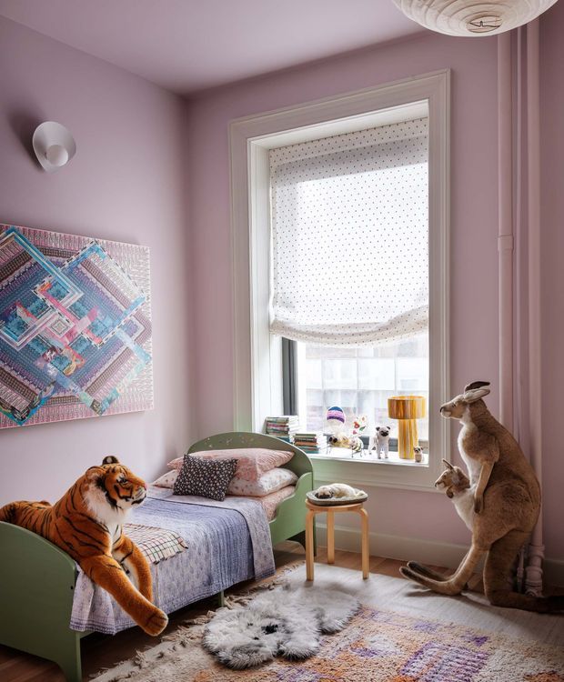 A playful, richly styled children's bedroom with dusty pink walls, a white-trimmed window and patterned Roman blind, a green-painted bed, layered pastel bedding, a small side table, and a pale textured rug over a patterned floor rug. Large tiger and kangaroo soft toys create a whimsical animal-themed atmosphere. A colorful geometric artwork and sculptural wall and ceiling lights add decorative interest.