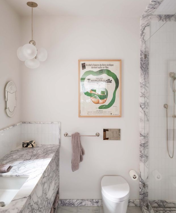 Refined bathroom interior featuring a freestanding or built-in bathtub with dramatic veined marble surround, a tiled shower enclosure with matching marble trim, a modern white toilet, brass-toned fittings, a globe pendant light, and a framed vintage poster. The space has a soft neutral palette and classic-modern detailing.
