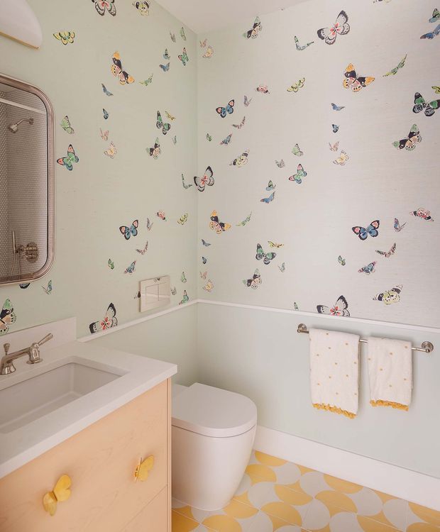 A compact, playful bathroom with pale mint-green walls covered in scattered butterfly wallpaper, white trim, and a light peach vanity with butterfly-shaped handles. The room includes a white basin, toilet, chrome fixtures, a rounded vintage-style mirror, patterned yellow-and-white floor tiles, and neatly hung towels with yellow trim.