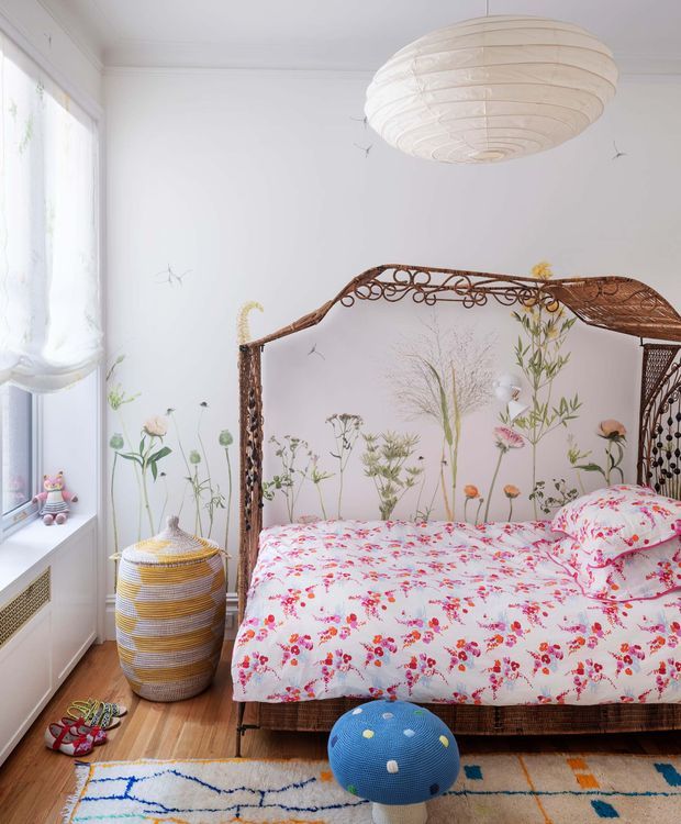 Bright, playful bedroom with a vintage woven metal canopy bed, white walls, floral botanical bedding, and a large white pendant light. The room features timber flooring, a patterned area rug, a blue round ottoman, a woven striped basket, tall sheer curtains, and botanical wall artwork. The overall atmosphere is colourful, cheerful, and eclectic with a soft natural palette.