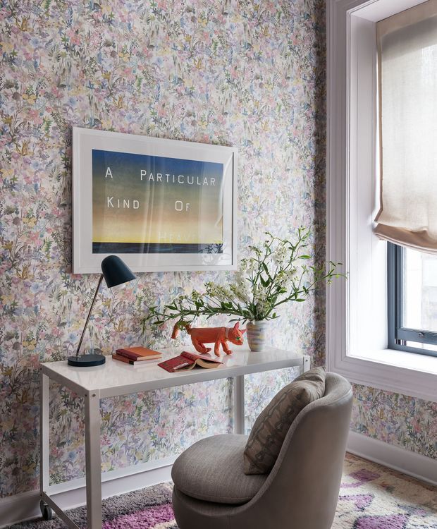 A compact, styled reading or work corner with a narrow white console desk against densely patterned floral wallpaper. The space includes a black angled desk lamp, books, framed artwork, a vase of leafy branches, and a small sculptural orange animal. A curved upholstered armchair sits in front of the desk on a multicoloured patterned rug. A large window with a light Roman shade provides natural light.