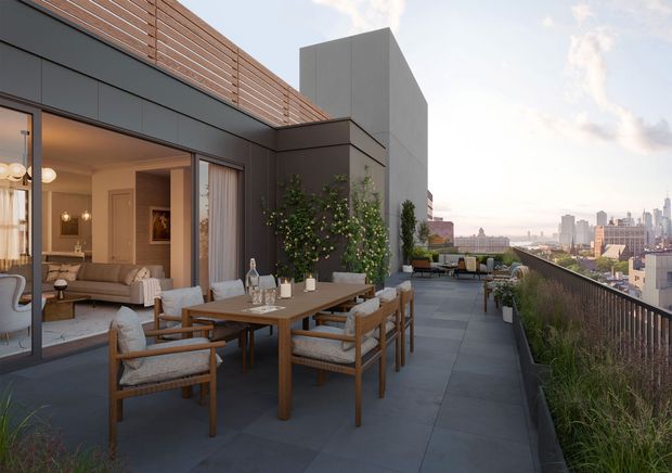 A rooftop terrace with a furnished outdoor dining area overlooking a dense city skyline. The terrace features large-format dark paving, a timber dining table with upholstered armchairs, planted borders, a dark metal balustrade, and a contemporary apartment facade with floor-to-ceiling glazing and timber privacy screening. A warm-toned living room is visible through the open doors.