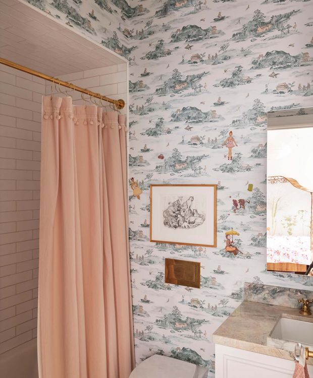 A compact, highly styled bathroom with a walk-in shower enclosed by pale pink subway tile and a blush shower curtain on a brass rod. The walls and ceiling feature whimsical illustrated toile-style wallpaper in soft pink, sage, grey, and cream. A framed artwork hangs above a brass flush plate, while a marble-look vanity benchtop, white inset basin, brass tapware, and a glimpse of a bedroom or adjoining space through the opening complete the scene.