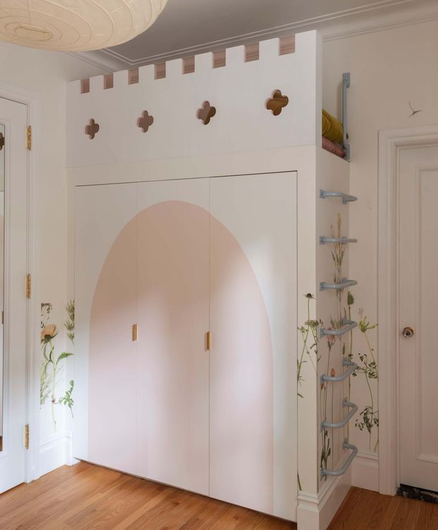 A whimsical built-in storage wall in a softly styled interior, featuring tall pale cabinetry with a large blush-pink arched inset, small brass pulls, decorative flower-shaped accents, and a crenellated upper edge. Open shelving with curved metal supports and trailing plants sits beside the joinery. The room has warm timber flooring, cream-painted walls and doors, decorative ceiling moulding, and a large sculptural pendant light.