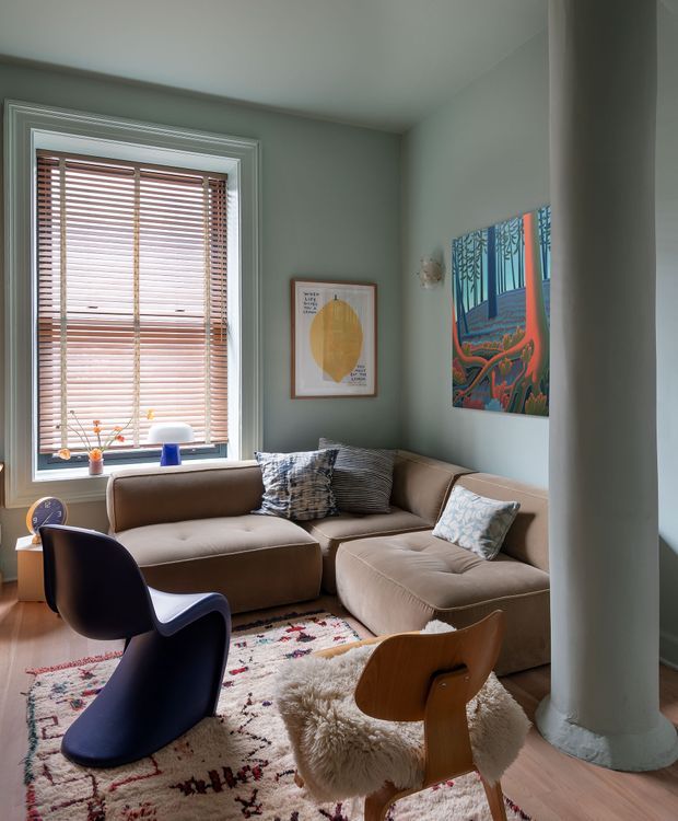 A compact, warmly styled living room with muted sage-grey walls, timber flooring, and a large bay-style window fitted with timber venetian blinds. A taupe sectional sofa wraps around the corner, accompanied by sculptural dark and timber lounge chairs, layered patterned and sheepskin rugs, and colourful framed artwork. A prominent round structural column stands at the right side of the room. The interior has a cosy, eclectic mid-century character with soft natural light.