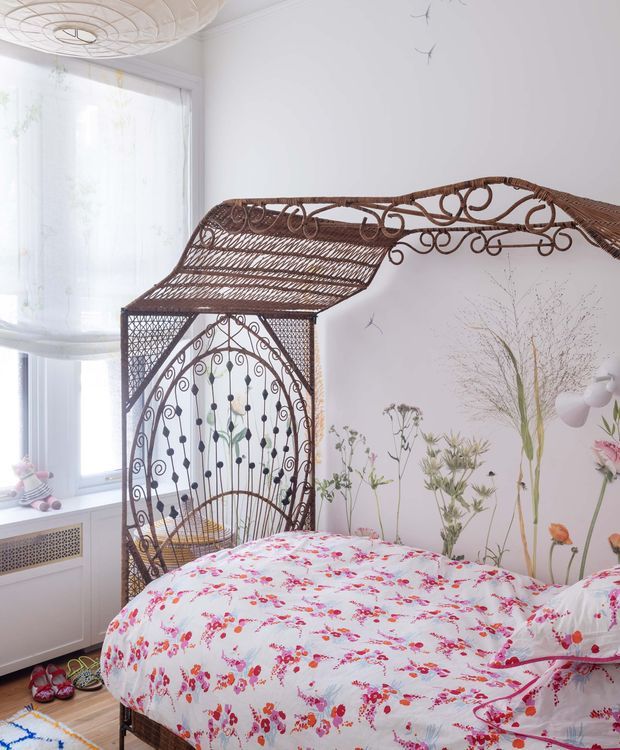 A bright bedroom with a vintage wrought-iron daybed or bed frame featuring elaborate scrolling and latticework canopy details. The bed is dressed in a white floral duvet, set against white walls with a delicate botanical mural or painted floral artwork. A large sheer-curtained window provides natural light, while a sculptural white ceiling light, radiator cover, and children's shoes add personal details. The overall look is whimsical, romantic, and eclectic.