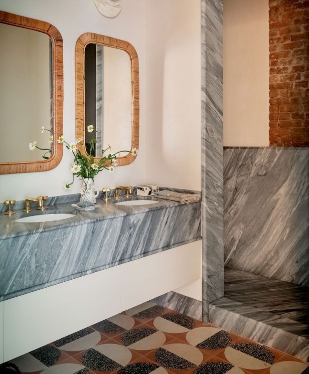 A refined bathroom featuring a double vanity with an expansive grey-veined marble benchtop and integrated basins, brass tapware, and rounded rattan-framed mirrors. The room includes dramatic grey marble wall and floor surfaces, an exposed red-brick wall section, pale plaster walls, and a patterned geometric tile floor in warm terracotta, cream, and dark tones. A floral arrangement adds a soft decorative accent.