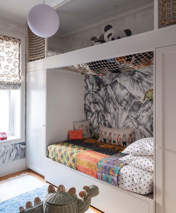 A playful children's bedroom featuring a built-in white bunk-style sleeping nook with integrated upper storage or play space, rope netting, and a patterned mountain mural wallpaper. The lower bed is dressed in a colourful patchwork quilt and whimsical animal cushions, with a woven rattan animal-shaped piece in the foreground. Natural light enters through a patterned roller blind, and the room has decorative ceiling moulding and a pale patterned wall finish.