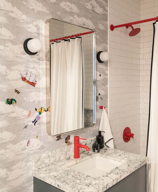 Playful, eclectic bathroom vanity and shower area featuring a marble-look benchtop with integrated white basin, grey vanity cabinetry, a rounded rectangular mirror, red faucet and shower fittings, black-and-white globe sconces, and contrasting wall finishes. The left wall has whimsical illustrated wallpaper with clouds and colorful figures, while the shower walls are clad in small white rectangular tiles with light grout. A white shower curtain and towels complete the compact, characterful space.