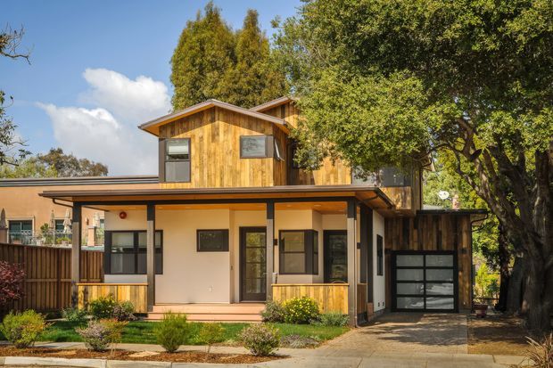 Exterior view of a contemporary multi-level home with warm timber cladding, light rendered walls, dark-framed windows, a broad low-pitched roof, and an integrated single-car garage. The upper storey projects over the ground-level entry and porch, while mature trees and landscaped planting frame the driveway.