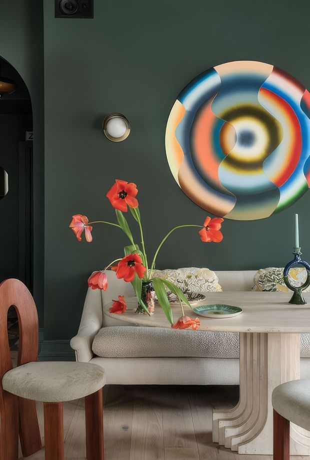 A richly styled dining or living area with a cream upholstered sofa or banquette, curved light-wood dining table, and matching timber chairs. The deep green wall features a large colourful abstract circular artwork and a small wall light. Red poppies in a vase, decorative objects, and patterned cushions add vivid accents. The space has a refined, eclectic, gallery-like atmosphere with warm natural timber and soft neutral upholstery.