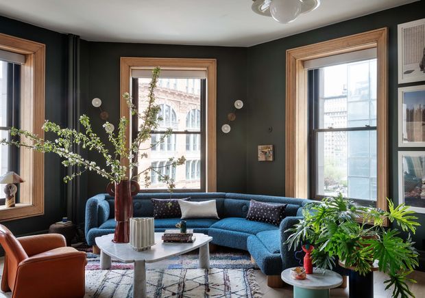 A richly styled living room with three large timber-framed windows, deep charcoal walls, and abundant natural light. A blue sectional sofa and rust-colored armchairs surround a sculptural white coffee table, with patterned cushions, a geometric rug, framed artwork, and large leafy indoor plants. The space has a refined, eclectic character with strong contrast between dark walls, warm timber, and vibrant upholstery.