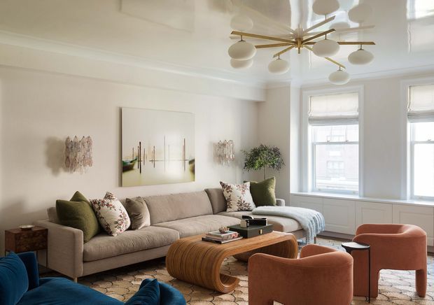 A bright, refined living room with a large neutral upholstered sofa, patterned and olive cushions, a sculptural rounded wood coffee table, rust-coloured lounge chairs, and a textured patterned rug. White walls, detailed ceiling mouldings, tall windows with Roman blinds, and a striking brass-and-glass globe chandelier define the room. Abstract artwork and plants add decorative interest.