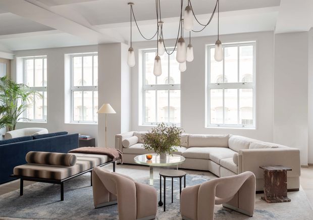 Bright, spacious contemporary living room with tall multi-pane windows, pale walls, a coffered ceiling, and sculptural pendant lights suspended at varying heights. The room is furnished with a large light sectional sofa, deep blue sofa, curved upholstered lounge chairs, a glass coffee table, a dark bench, and a textured area rug. Neutral tones are complemented by blue upholstery, greenery, and warm wood accents.