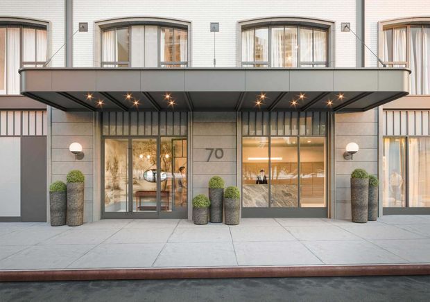 Street-level view of a refined multi-storey building entrance with a broad dark metal canopy, recessed star-like downlights, pale masonry facade, tall glazed doors and windows, and the number 70 displayed beside the entry. Symmetrical cylindrical planters with clipped spherical shrubs frame the entrance, while warm interior lighting and reception-like furnishings are visible through the glazing.