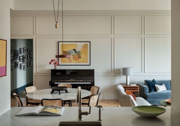 A refined open-plan interior viewed from a kitchen island, with a white paneled wall, black upright piano, round dining table and chairs, and a blue sofa seating area. The room features pale timber flooring, framed artwork, sculptural pendant lighting, and a light stone kitchen benchtop with a traditional-style faucet. The overall composition is bright, elegant, and layered with classic architectural detailing and contemporary furnishings.