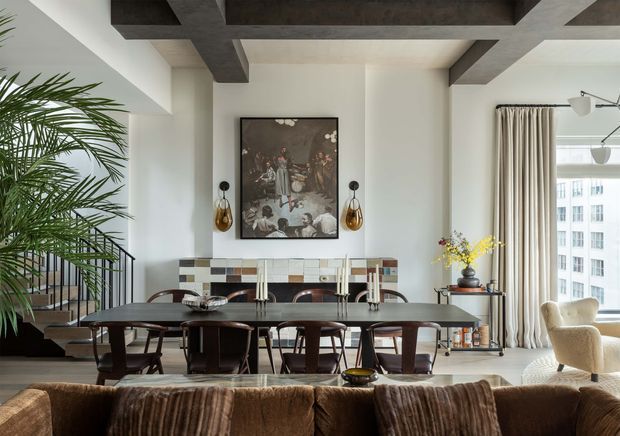 A refined open-plan dining and living area in an apartment, centred on a long dark timber dining table with sculptural dining chairs. The room features exposed dark-stained ceiling beams, white walls, a tiled fireplace or feature wall behind the table, contemporary artwork, brass pendant wall lights, full-height beige curtains, and large windows overlooking an urban setting. A sofa occupies the foreground, while a palm and stair railing are visible to the left.