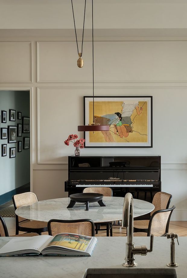 A refined dining area viewed from a kitchen island, featuring a round marble dining table with cane-back chairs, a black upright piano, framed artwork, decorative wall moulding, and a sculptural pendant light. A partial kitchen sink and chrome tap are visible in the foreground, while a gallery wall appears through the opening on the left.