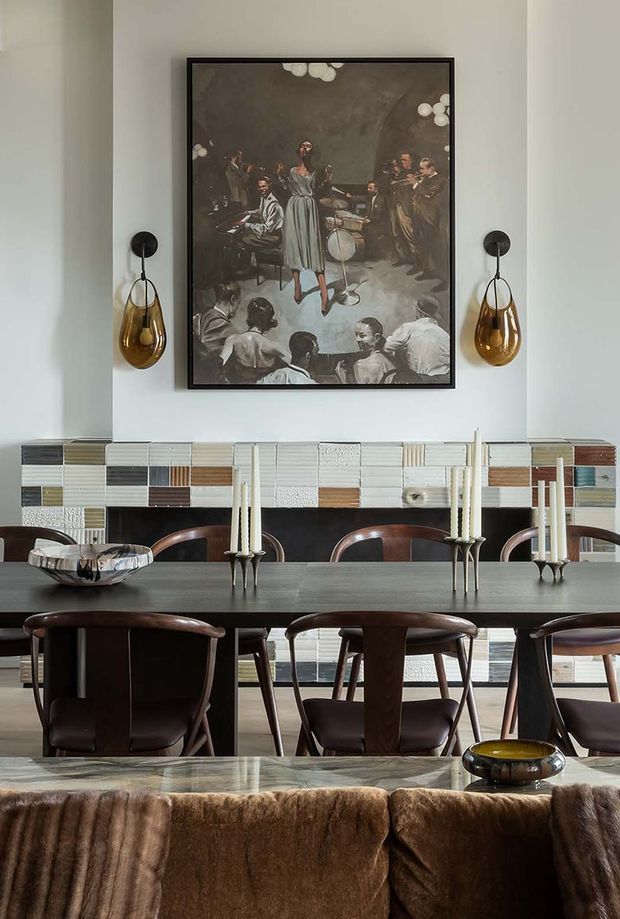 A refined dining room viewed from behind a sofa, featuring a long dark dining table with sculptural wooden chairs, multiple candlesticks, a large framed artwork, pendant wall sconces, and a striking multicoloured tiled feature wall. The space has a moody, richly layered palette with dark timber, warm amber glass, cream walls, and textured finishes.