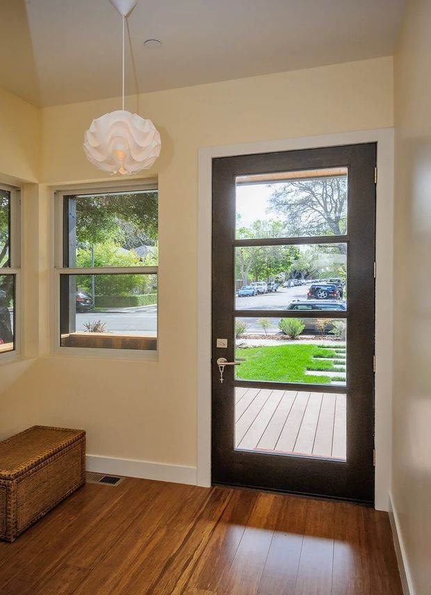 Bright residential entryway with cream-painted walls, warm timber flooring, a dark framed glazed exterior door, adjacent windows, and a sculptural white pendant light. The door opens toward a small timber deck and landscaped front garden, with a woven storage chest positioned along the wall.