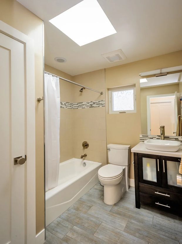A bright, compact bathroom with a bathtub and shower curtain, toilet, white vessel sink, and dark wood vanity. Beige walls are paired with a horizontal mosaic tile accent band, grey wood-look floor tiles, a frosted window, and a large rectangular skylight.