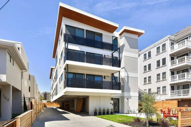 Exterior view of a contemporary multi-storey residential building with white rendered walls, grey horizontal cladding, broad projecting roof eaves, extensive dark-framed glazing, and multiple balconies with black horizontal metal balustrades. The building is raised over a driveway or lower-level parking area, with neighbouring residential buildings visible on either side and a small landscaped frontage.