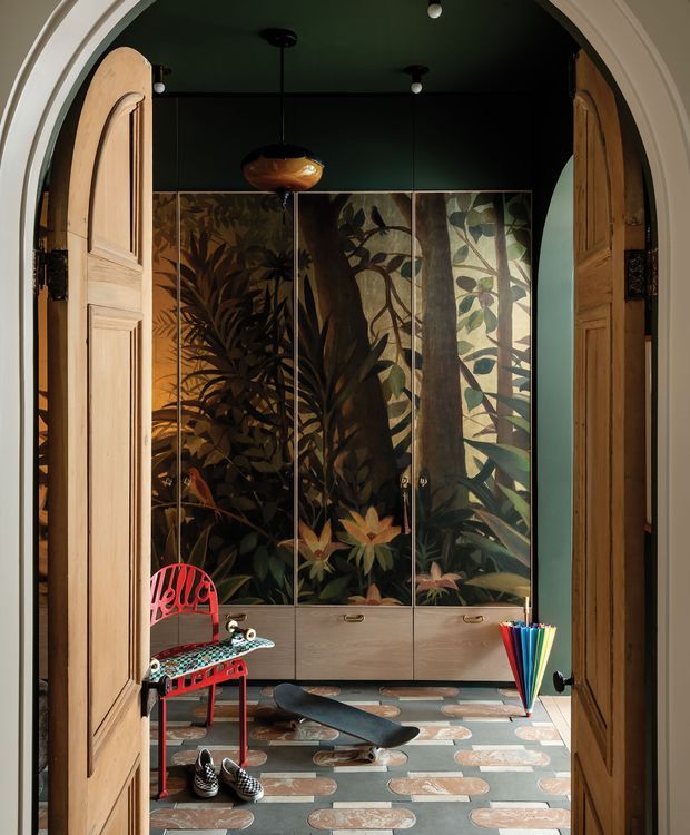 A moody, eclectic entryway viewed through open arched timber double doors. The space has a deep green ceiling and walls, a large botanical mural across built-in cabinetry, patterned stone or tile flooring, a sculptural pendant light, and playful red furniture with a skateboard and colourful umbrella. The composition combines traditional architectural detailing with bold artistic and contemporary furnishings.