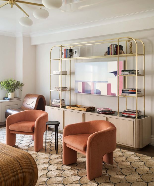 A refined living room with two sculptural rust-toned lounge chairs and a matching curved chair arranged on a patterned carpet. A light timber built-in cabinet and open brass-framed shelving occupy the rear wall, displaying books, ornaments, and abstract artwork. The room has cream walls with decorative ceiling cornices, a contemporary brass multi-globe pendant, and a warm, elegant palette.