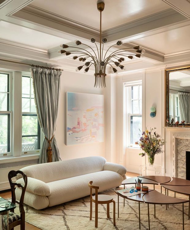 Elegant, light-filled living room with high decorative coffered ceilings, layered crown moulding, tall black-framed windows, and a carved fireplace mantel with mirror. A curved cream sofa, sculptural vintage-inspired chandelier, nesting wood coffee tables, patterned rug, and restrained artwork create a refined eclectic interior.