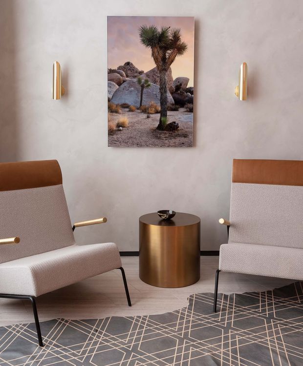A refined seating area with two mid-century-inspired upholstered lounge chairs featuring warm brown leather headrests, pale textured fabric, slim black legs, and brass-toned arm details. A cylindrical bronze side table sits between them beneath a desert landscape artwork, flanked by sculptural brass wall sconces. The space has a softly textured light plaster wall, pale timber flooring, and a geometric dark rug.