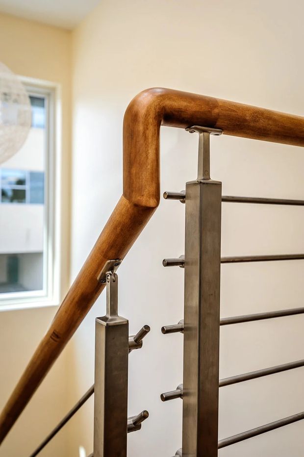 Close-up interior view of a contemporary stairway railing. The handrail is made of warm stained timber with rounded, sculpted joints, supported by brushed metal vertical posts and horizontal metal balusters. A pale neutral wall and window form the background, with a partial spherical pendant light visible at the left.