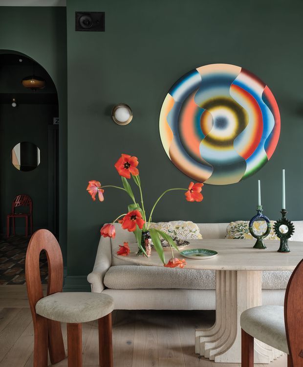 A richly styled dining and living area with a dark green feature wall, sculptural multicoloured circular wall art, cream upholstered sofa, pale stone or painted timber dining table, and curved timber dining chairs. Decorative candles, glassware and a vibrant floral arrangement add colour. An arched opening leads to a darker adjoining space, while the room features warm timber flooring and layered, eclectic furnishings.