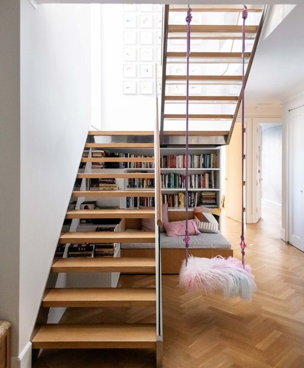 Bright contemporary interior featuring an open timber staircase with slim dark metal supports and integrated open shelving filled with books beneath and behind the stairs. A playful suspended swing hangs beside the staircase, overlooking a cosy reading nook with a sofa and cushions. The room has white walls, pale timber parquet flooring, abundant daylight, and framed artwork on the upper wall.