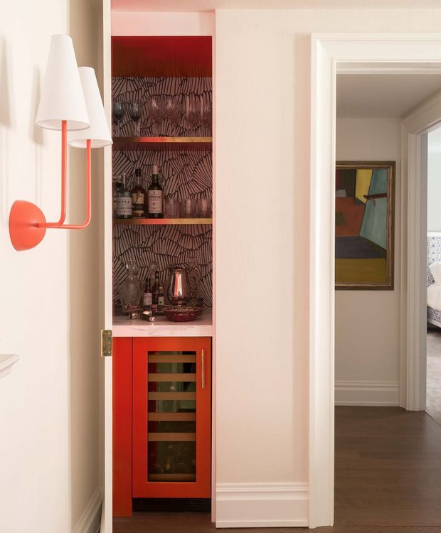 A refined residential hallway or living-area view featuring a built-in red home bar with a glass-front wine refrigerator, open bottle shelving, and a patterned dark backsplash. A sculptural white-and-coral wall sconce is mounted beside the joinery. Through the doorway, a framed artwork and part of a bedroom with patterned bedding are visible. The space has white painted walls, substantial white door trim and skirting, and dark timber flooring.