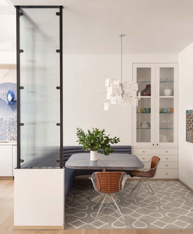 Bright, refined dining area with a mid-century-inspired table and sculptural wire-frame chairs, arranged beside a built-in upholstered banquette. A tall black-framed ribbed glass screen partially divides the space, while a white built-in display cabinet with glass doors provides storage. The room features a patterned grey area rug, pale walls and timber flooring, a suspended paper-note style pendant, and a leafy plant centerpiece.