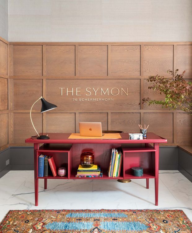 A styled home office or study vignette featuring a vivid red/pink writing desk with open shelving, books, a laptop, desk lamp and accessories. The desk is positioned against a large warm timber-panelled feature wall with geometric framed sections. Pale marble-look flooring and a richly patterned oriental rug complete the room, with a leafy indoor plant at the side. The image includes promotional text reading “THE SYMON 76 SCHERMERHORN.”