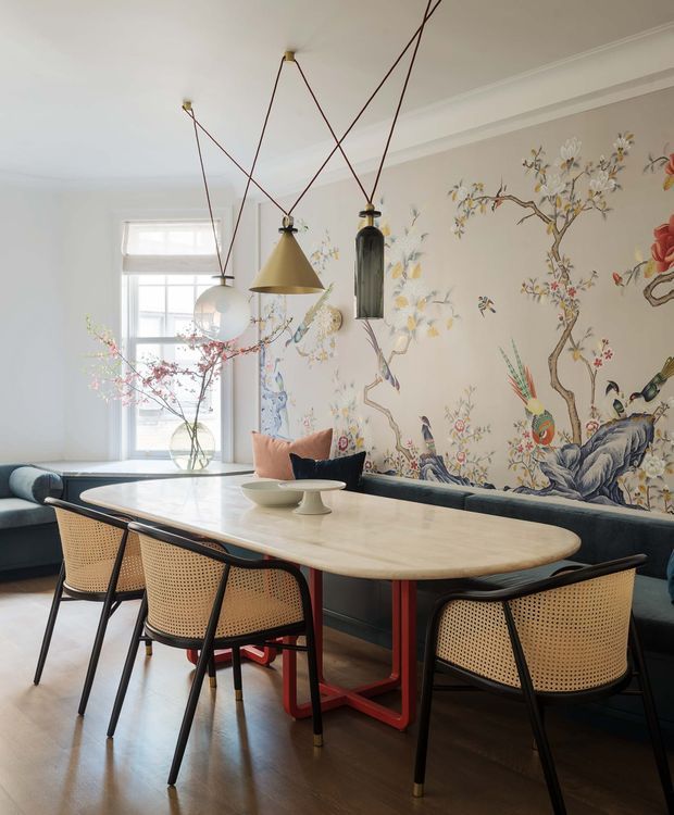 Bright, refined dining area with a large oval light-stone dining table, cane-back black-framed chairs, and a built-in dark banquette. A dramatic botanical and chinoiserie-style mural covers the feature wall, complemented by sculptural pendant lights with exposed dark cords. Tall glazing and pale walls create an airy setting, with dark timber flooring and floral branches adding warmth.