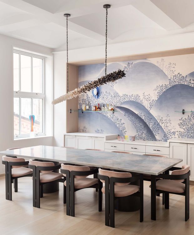 A bright, refined dining room with a large rectangular dining table and multiple upholstered chairs. A built-in white sideboard and open glass shelving sit against a dramatic blue and lavender mural, while a sculptural linear pendant hangs above the table. Tall windows provide abundant natural light, and the ceiling features subtle recessed geometric detailing.