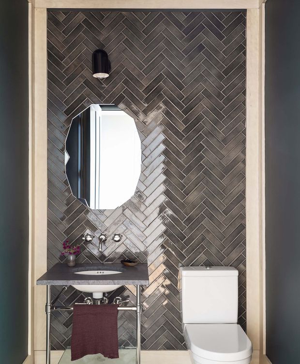 Compact powder room with a dramatic dark grey herringbone tile feature wall, framed by pale trim. The room includes a wall-mounted oval mirror, black pendant light, stone vanity basin with exposed chrome plumbing, small floral arrangement, and a white toilet. The design is refined and high-contrast, with a strongly patterned tiled backdrop.