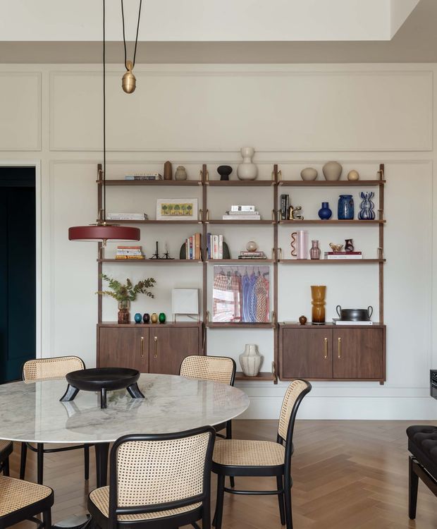 Refined dining room with a large round marble dining table surrounded by cane-back chairs, a dark sculptural centerpiece, and a wall-length built-in shelving and display unit. The joinery is dark timber with brass-toned hardware and open shelves displaying ceramics, books, glassware, and artwork. White decorative wall panelling, a suspended red pendant light, herringbone timber flooring, and a dark doorway complete the elegant interior.