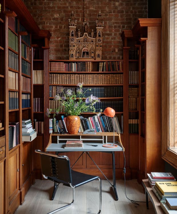 A compact, richly detailed home library and study with floor-to-ceiling dark timber bookshelves filled with books, a small modern desk, and a black leather-and-chrome chair. The room has exposed red brick walls, pale timber flooring, warm natural light from a tall timber-framed window, and eclectic decorative objects including a carved architectural ornament, vase of flowers, and orange desk lamp. The overall character is scholarly, warm, and highly layered, combining traditional built-in joinery with mid-century furniture.