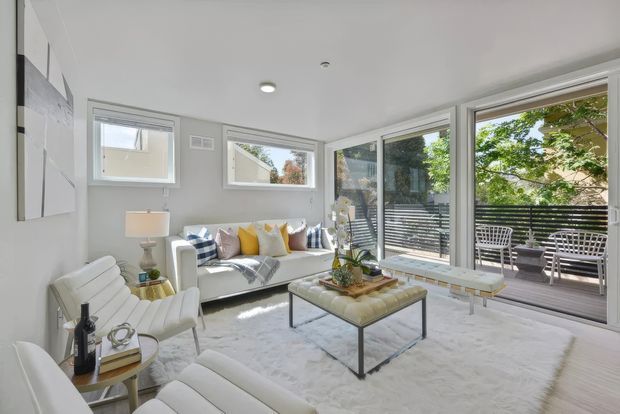 Bright, airy contemporary living room with white upholstered lounge furniture, a pale area rug, glass-topped coffee table, and large corner glazing opening to a deck or terrace. Soft yellow and blue cushions add restrained colour accents, while the neutral finishes and abundant natural light create a clean, inviting atmosphere.